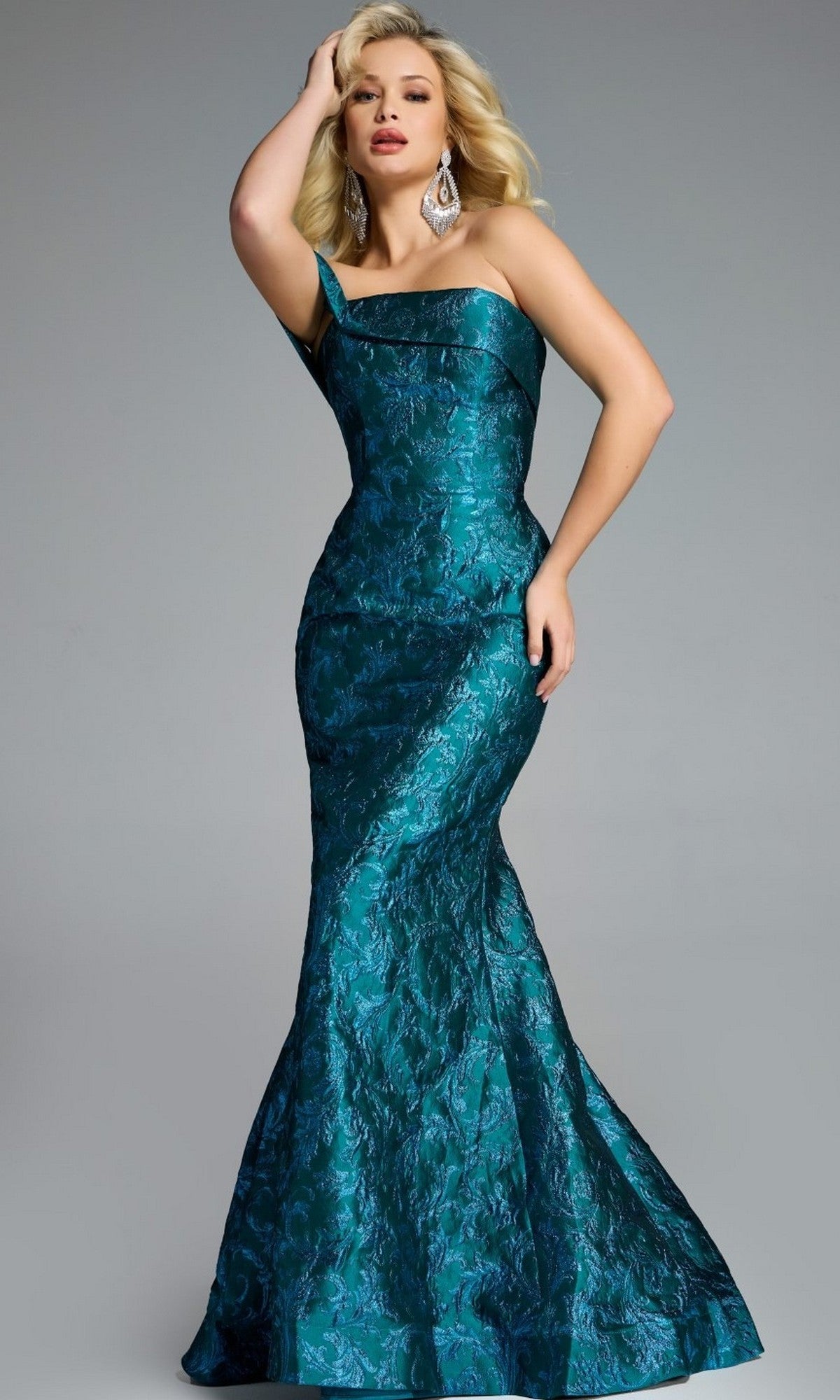 A woman poses in a fitted, teal Jovani 43978 long prom dress with a one-shoulder design and textured floral pattern against a plain gray background.